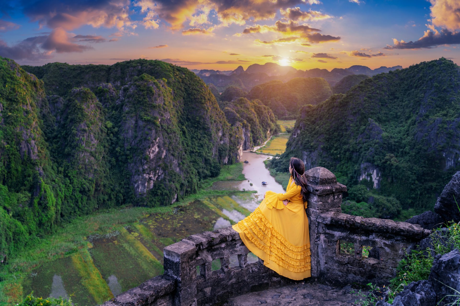 A woman in a flowing yellow dress looks out over the Tam Coc river valley from the Mua Cave viewpoint as the sun sets behind Ninh Binh's limestone peaks.
