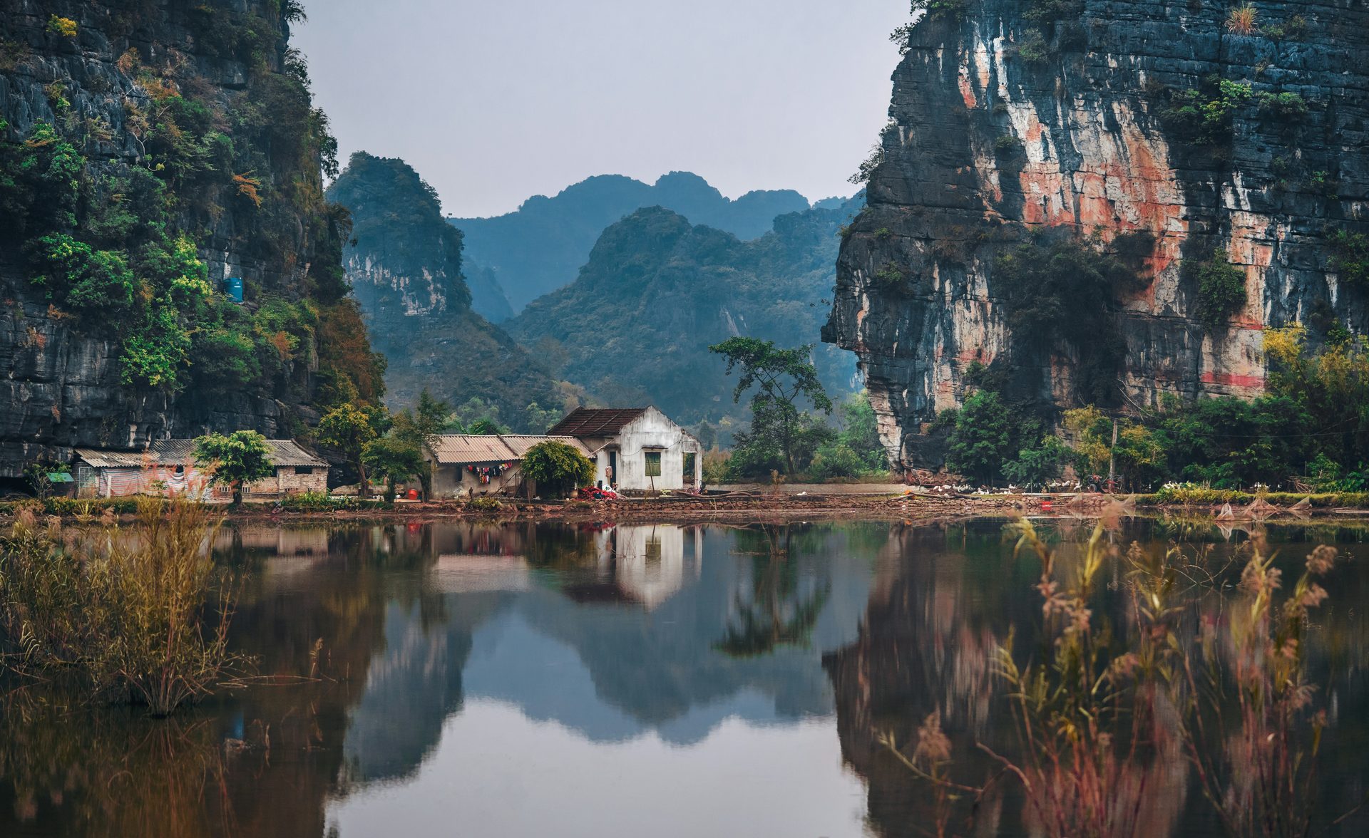 A small white house sits among limestone cliffs, its reflection mirrored in the still river water of the Ninh Binh countryside.