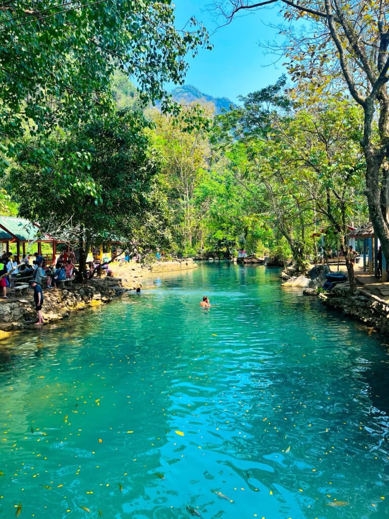 Turquoise natural swimming pool surrounded by forest and limestone cliffs near Vang Vieng, Laos, with visitors swimming