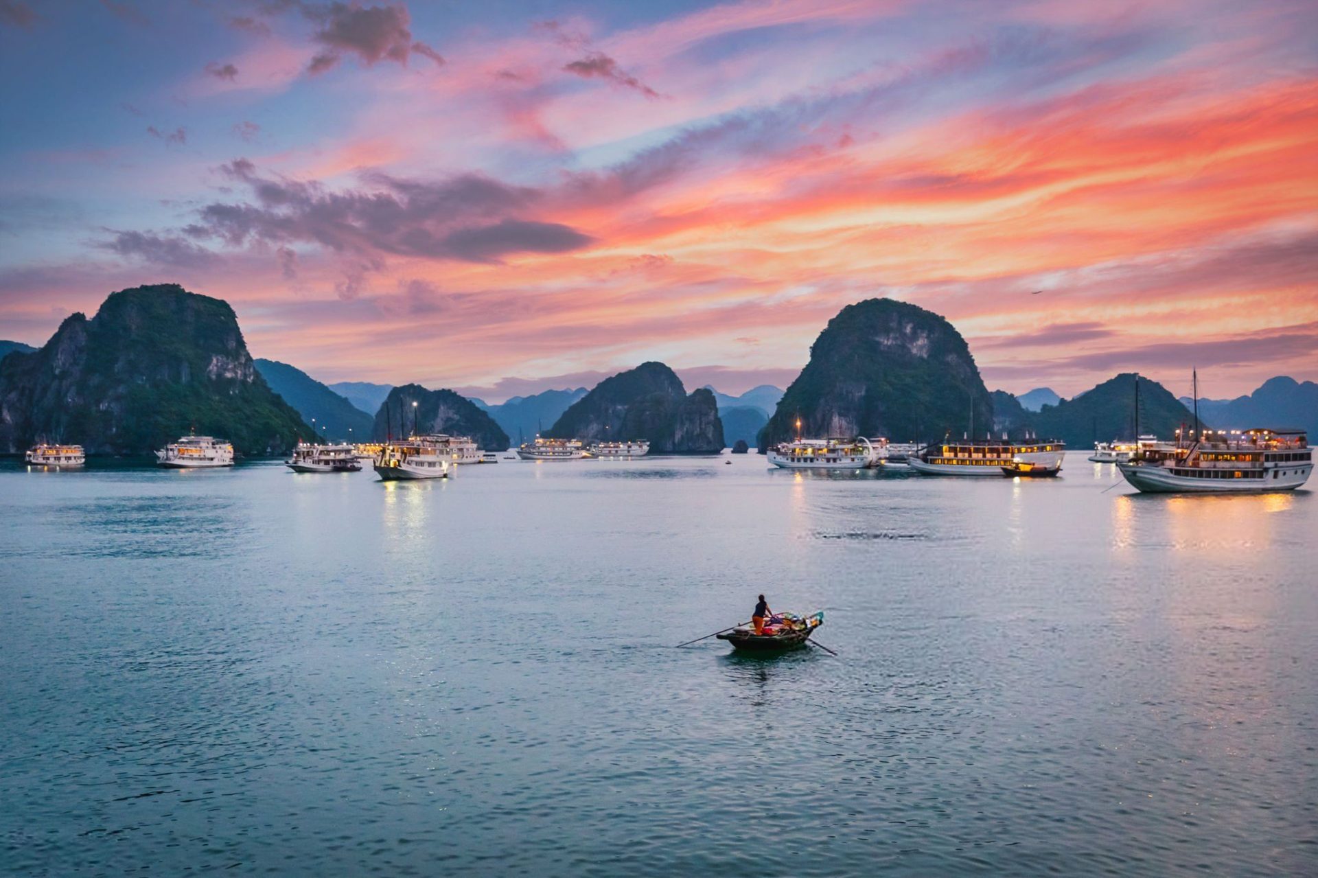 A wooden boat rows across Ha Long Bay as the sky turns pink and orange at sunset, with limestone islands and lit cruise boats in the distance.