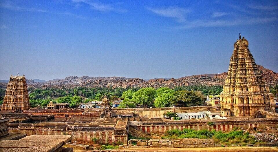 Panoramic view of Hampi's ancient ruins with Virupaksha Temple's gopuram rising above boulder-strewn hills and green groves.