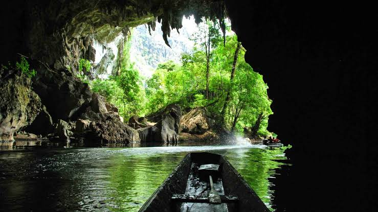 Wooden boat gliding through the flooded entrance of Kong Lor Cave beneath towering limestone karst, Laos