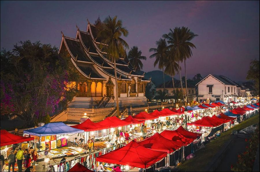 Evening stalls of the Luang Prabang Night Market lit up beneath palm trees with the former Royal Palace behind, Laos