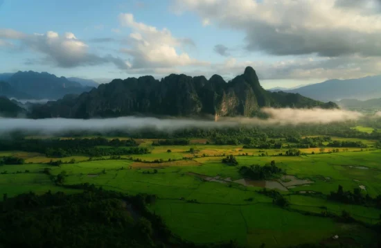Mist drifting over green rice fields beneath the limestone karst mountains of Vang Vieng, Laos