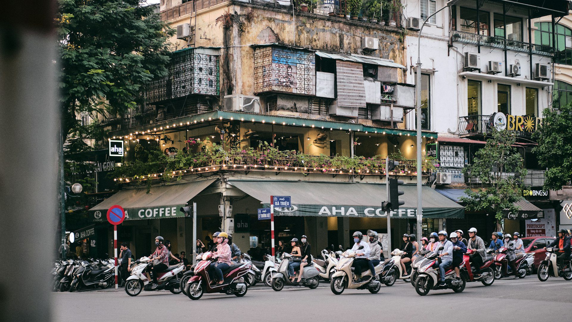 Motorbikes weave past narrow shophouses on a busy street in Hanoi's Old Quarter during the day.