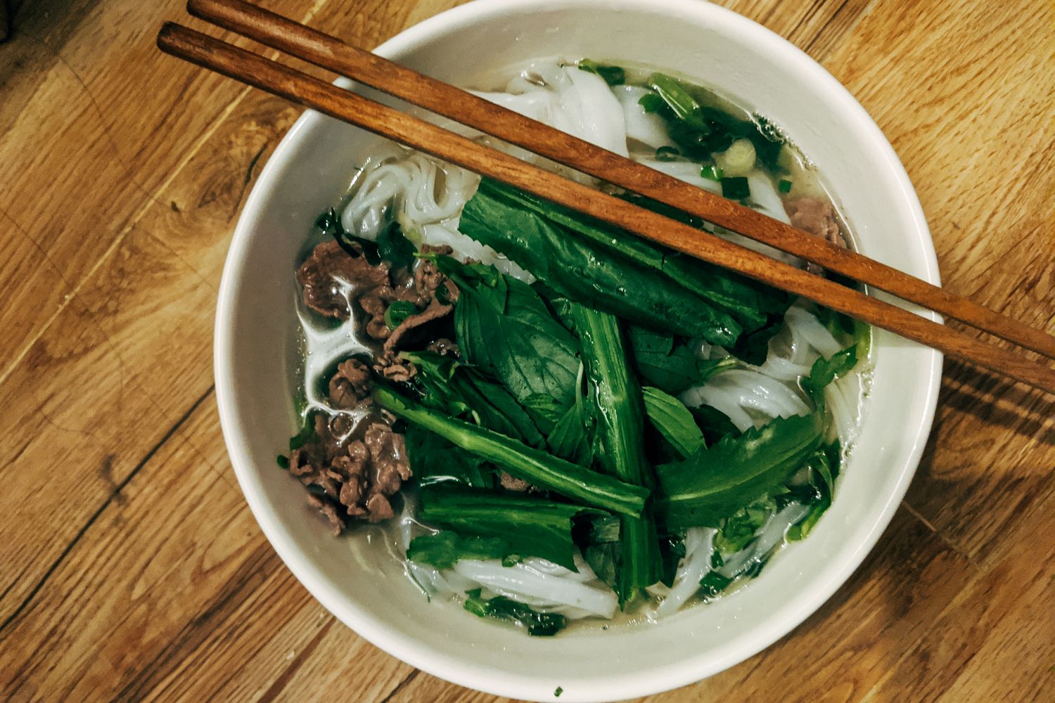 A bowl of Vietnamese pho topped with fresh herbs, served with wooden chopsticks on a wooden table.